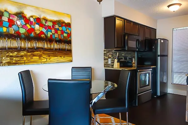 Glass-top dining table and chairs beside a modern kitchen with dark cabinets and stainless appliances.
