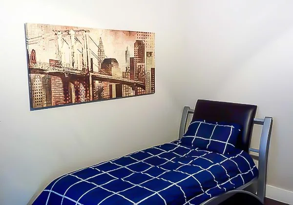 Single bed with blue bedding in a furnished bedroom with white walls and framed wall art.