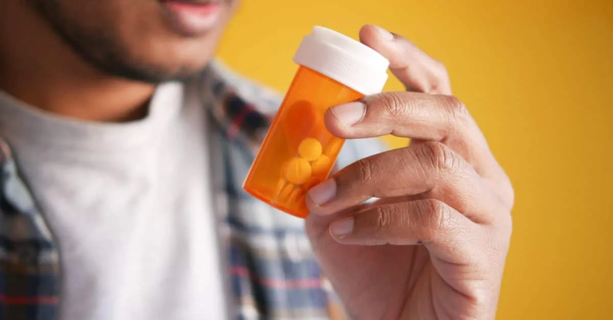 Close-up of a man holding a prescription medicine container against a yellow background.