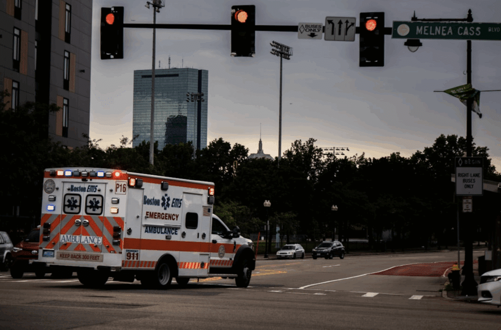 A Boston EMS ambulance driving through an intersection with emergency lights on under a cloudy evening sky.