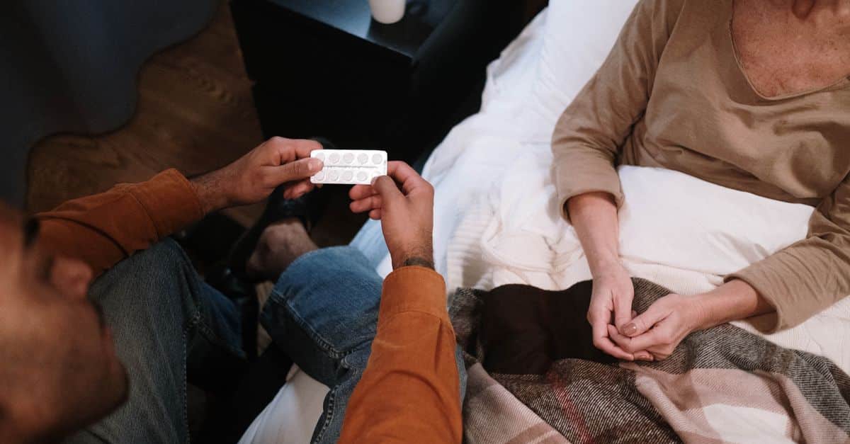 A caregiver sits next to a senior in bed, discussing medication in a home setting.