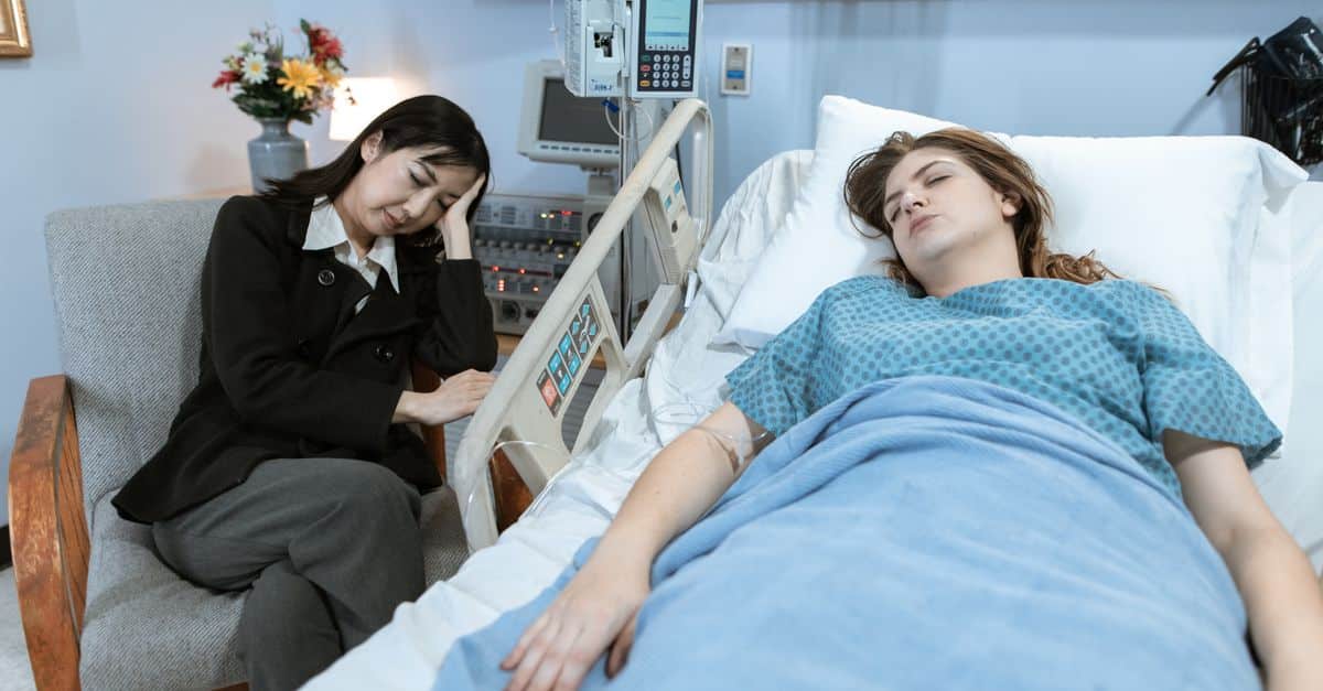 A woman sits by a hospital bed, looking worried, as a patient rests peacefully.