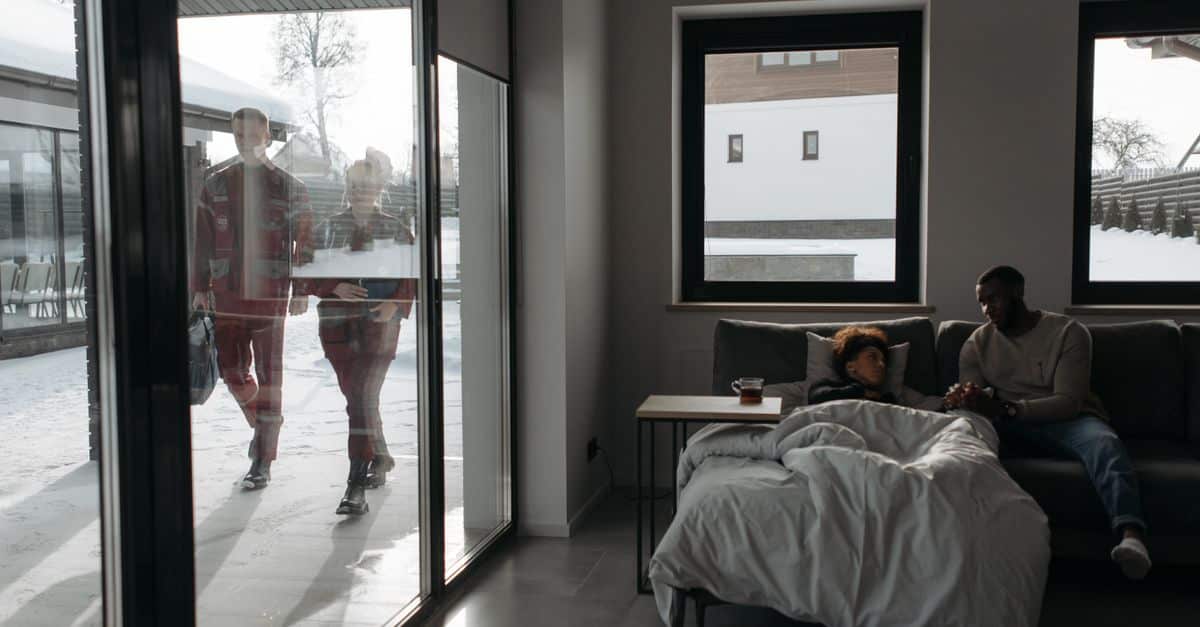 Healthcare professionals approach a patient's home through snow, providing emergency care indoors.