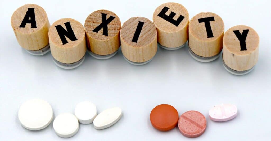 Wooden blocks spelling 'Anxiety' with assorted pills below on a light background.