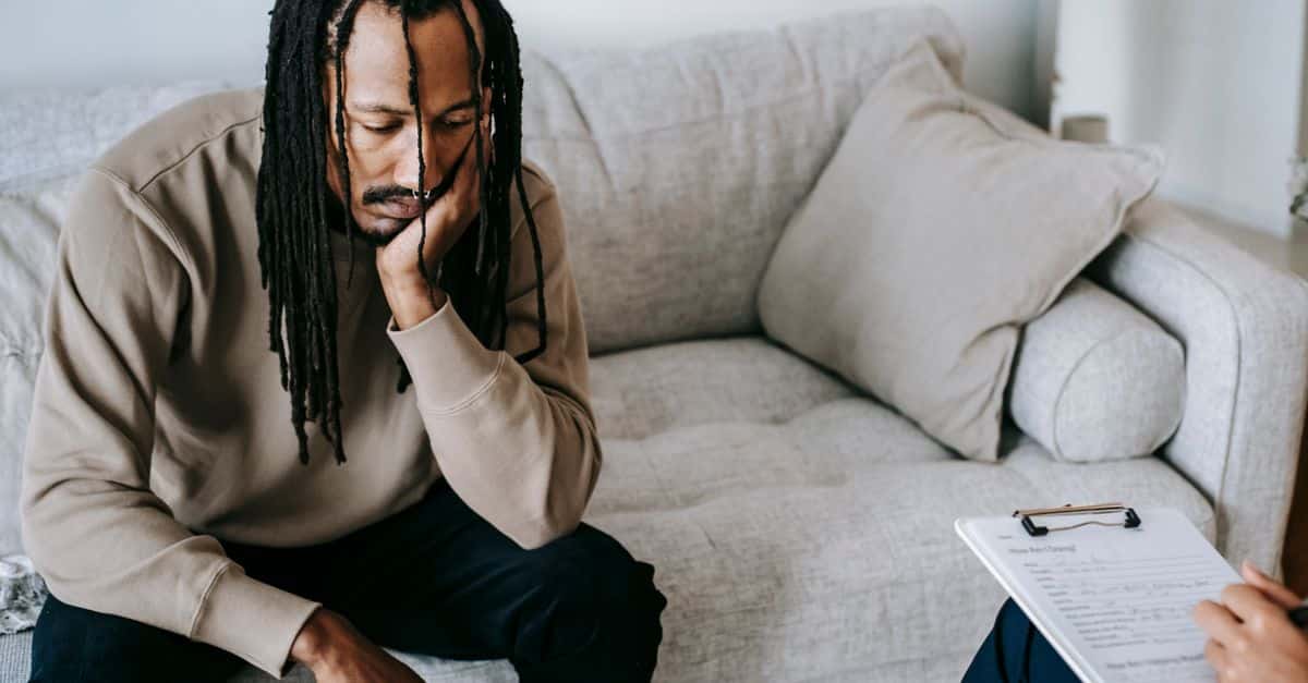 Upset African American male with dreadlocks leaning on hand and looking down while sitting on couch near crop psychologist with clipboard