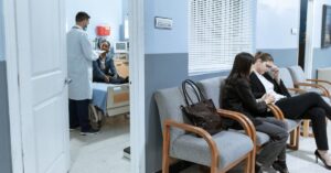 Doctor with patient in ICU, women in waiting area showing concern.