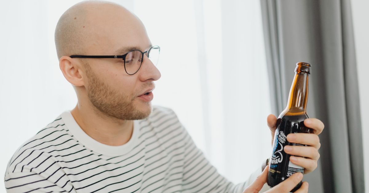 Bald man with glasses examines a bottle of craft beer indoors.