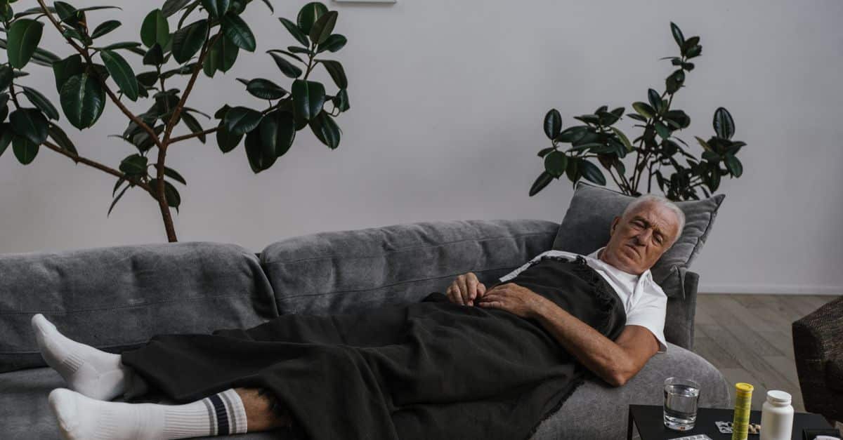 Elderly man resting on sofa with medical supplies nearby, surrounded by houseplants.