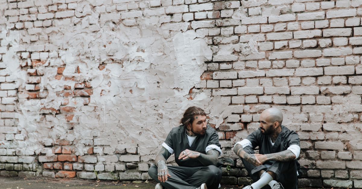 Tattooed men in prison uniforms talking while sitting outdoors against a brick wall.