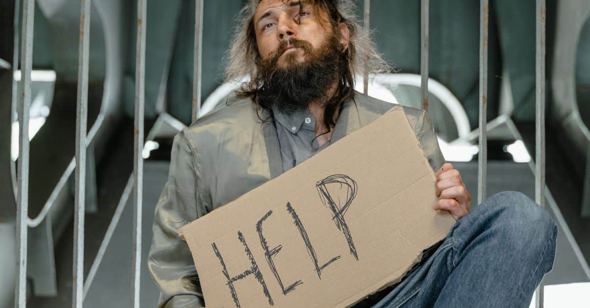 A homeless man with a beard holding a sign asking for help indoors.