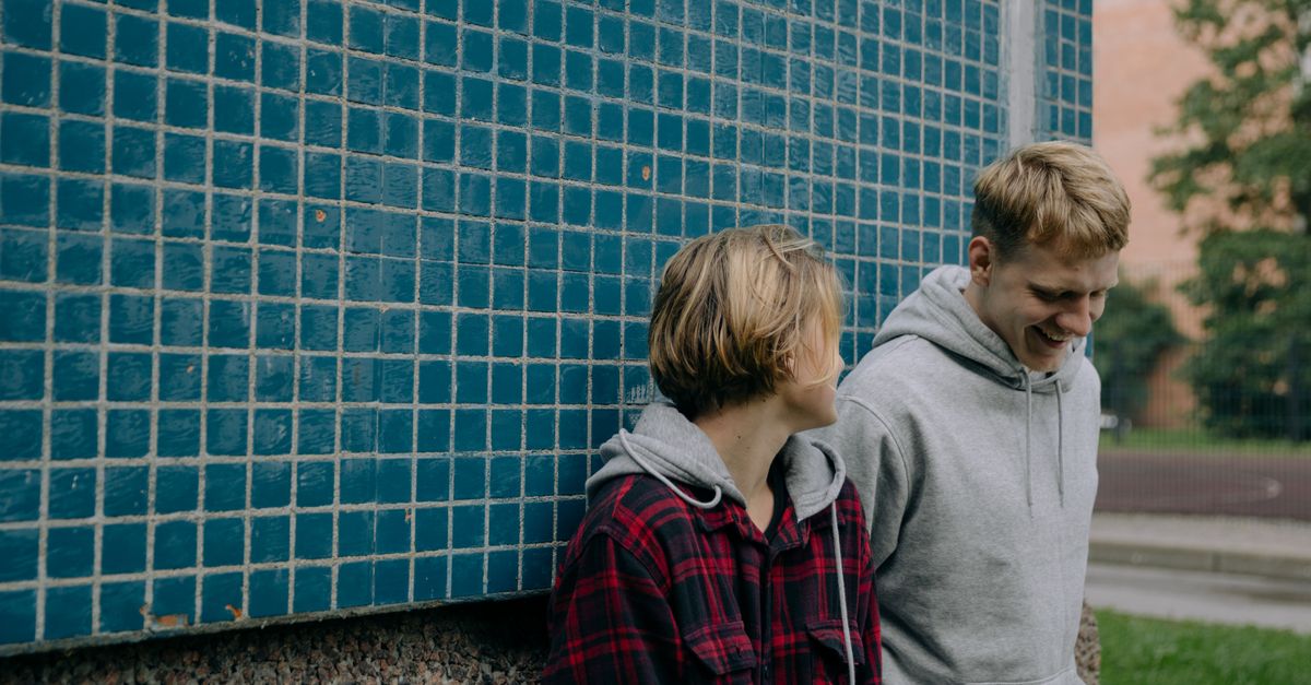 Two young friends in casual attire laughing against a blue tiled wall outdoors, enjoying a casual moment together.