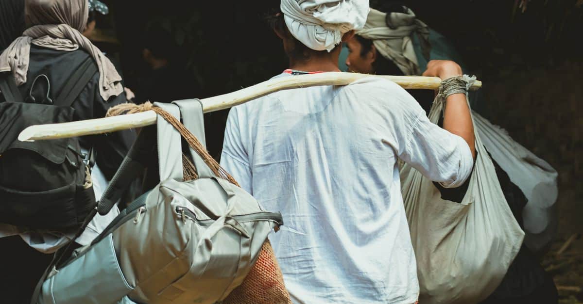 A traveler carrying multiple bags using a wooden pole, going through a rural setting.