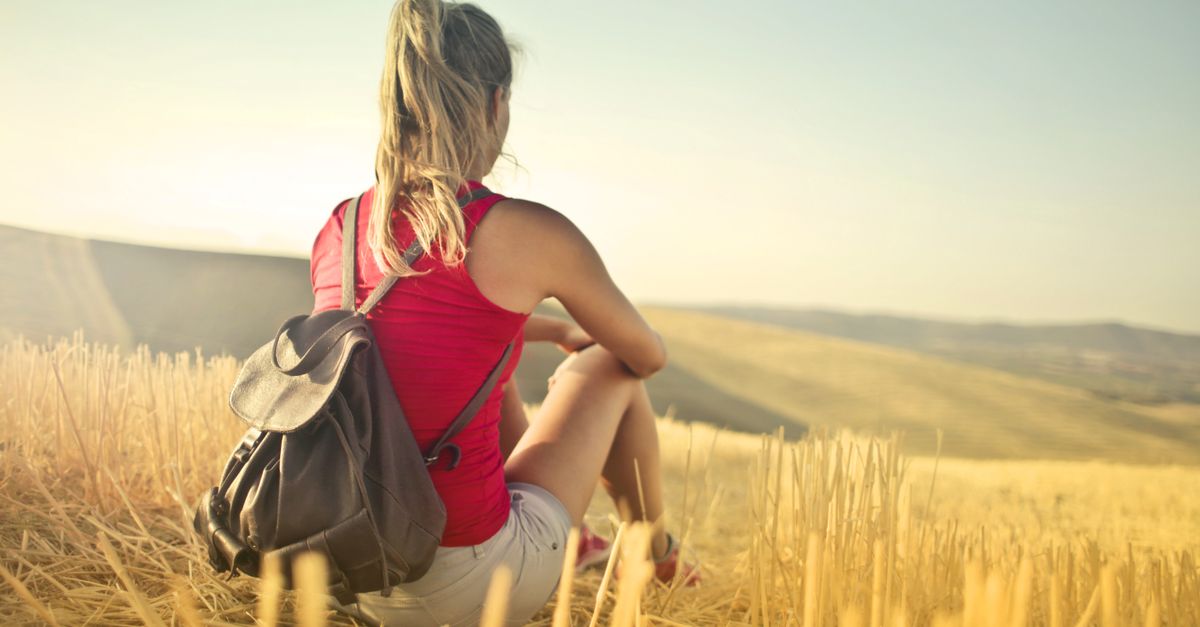 Woman with backpack sits in a golden hayfield, embracing solitude on a sunny day.
